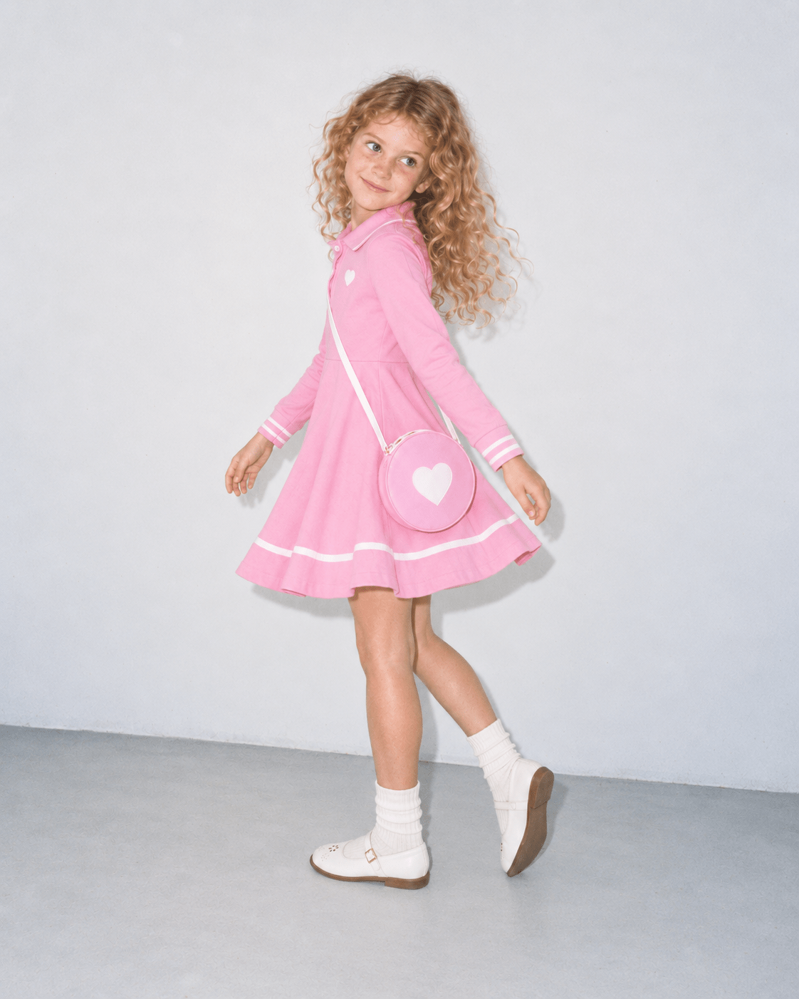Girl wearing a pink long sleeve polo dress while walking, showing the side view, flared hem and matching heart shoulder bag.