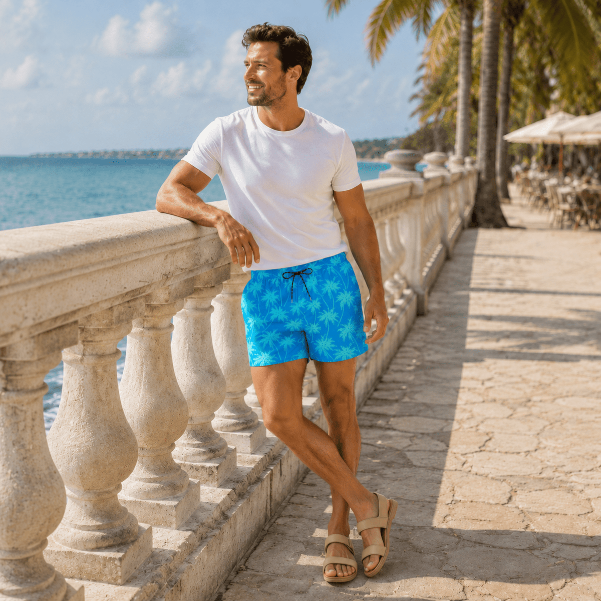Man wearing Blue Palm 5.5 Inch swim shorts with a white T-shirt and sandals on a sunny seaside resort walkway.