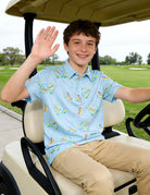 Boy sitting in a golf cart and waving while wearing a light blue short sleeve polo shirt with colorful golf cart prints and khaki pants.
