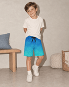 Boy wearing blue gradient palm tree swim trunks with a white T-shirt, showing the relaxed fit, tropical print and summer beach style.