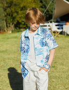 Boy standing on grass in a blue palm tree print button-down shirt layered over a white tee, with one hand in his pocket.