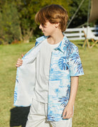 Boy wearing a blue palm tree short sleeve button-down shirt open over a white T-shirt, styled as a casual layered outdoor outfit.
