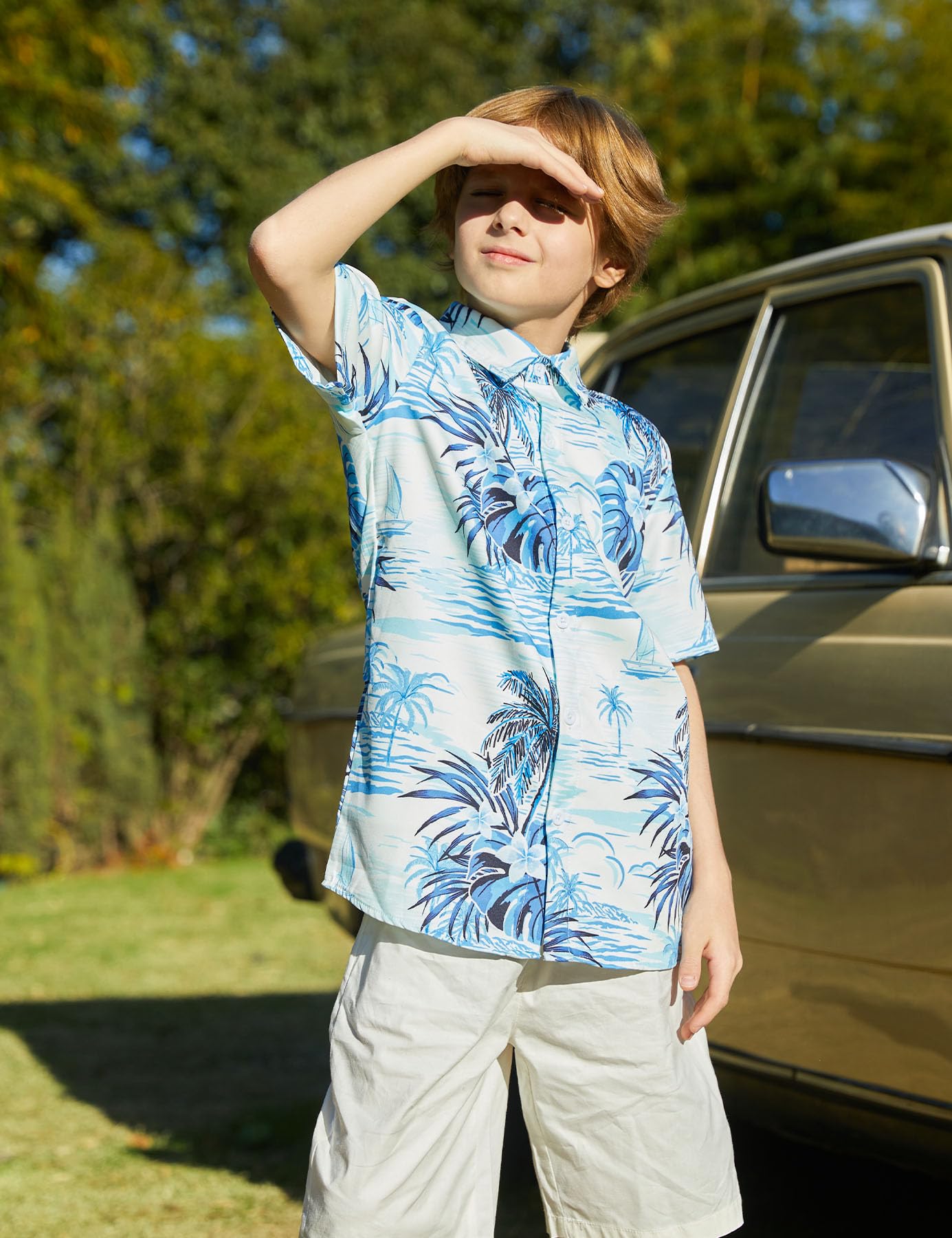 Boy wearing a light blue palm tree print button-down shirt outdoors beside a vehicle, raising his hand to shade his eyes from the sun.