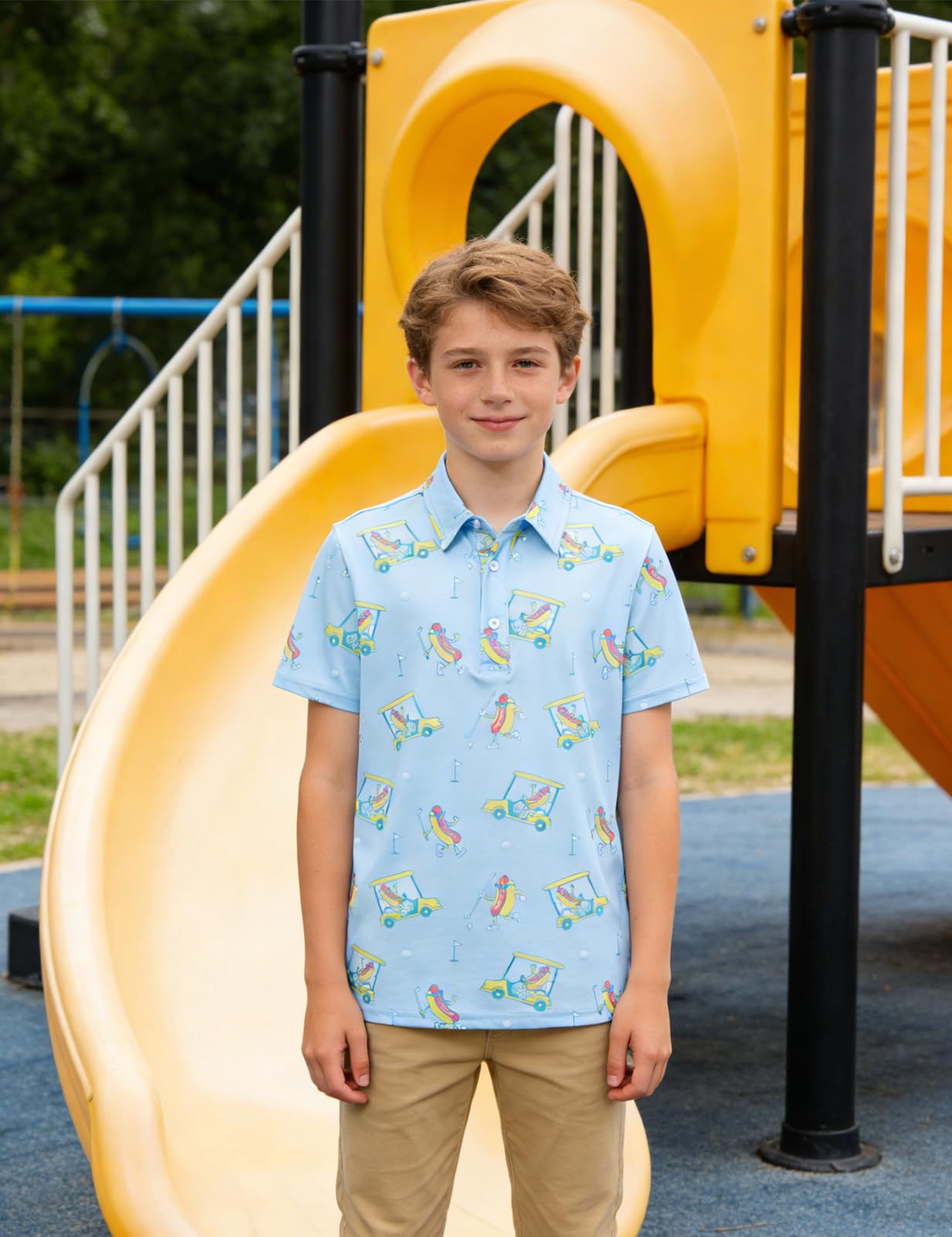 Boy standing in front of a yellow playground slide wearing a light blue short sleeve polo shirt with colorful golf cart prints and khaki pants.
