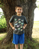 Boy wearing a dinosaur graphic tee with blue shorts, posing outdoors and showing a fun casual outfit for daily wear.