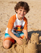 Boy wearing an Orange Hawaii rash guard swim set while building a sandcastle on the beach, styled for summer beach play.