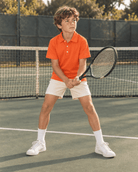Boy wearing an orange short sleeve polo shirt with shorts while standing on a tennis court, showing the comfortable everyday active fit.