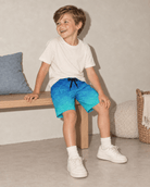 Boy sitting in blue gradient palm tree swim trunks, showing the comfortable fit and tropical design for beach trips, pool days and summer vacations.