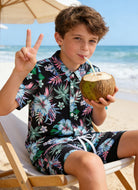 Boy wearing tropical printed swim shorts with matching shirt, lightweight summer clothing for beach trips and holidays.