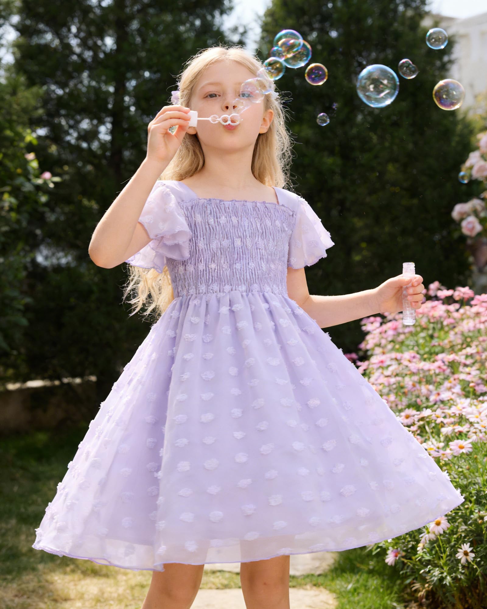 Girl wearing a lavender Swiss dot chiffon dress in a garden setting, showing the flowing skirt and elegant ruffle sleeve design.