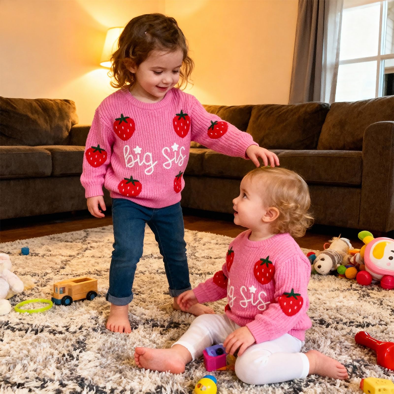 Two young sisters wearing pink sibling knit sweaters while playing together indoors, styled for cozy everyday and family moments.