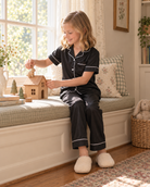 Girl wearing a black pajama set while sitting indoors, showing the relaxed fit, button front top and matching sleepwear pants.