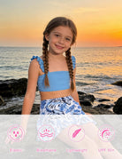 Girl wearing a blue 3 piece swim set with floral skirt by the ocean at sunset, showing a colorful summer beach outfit.