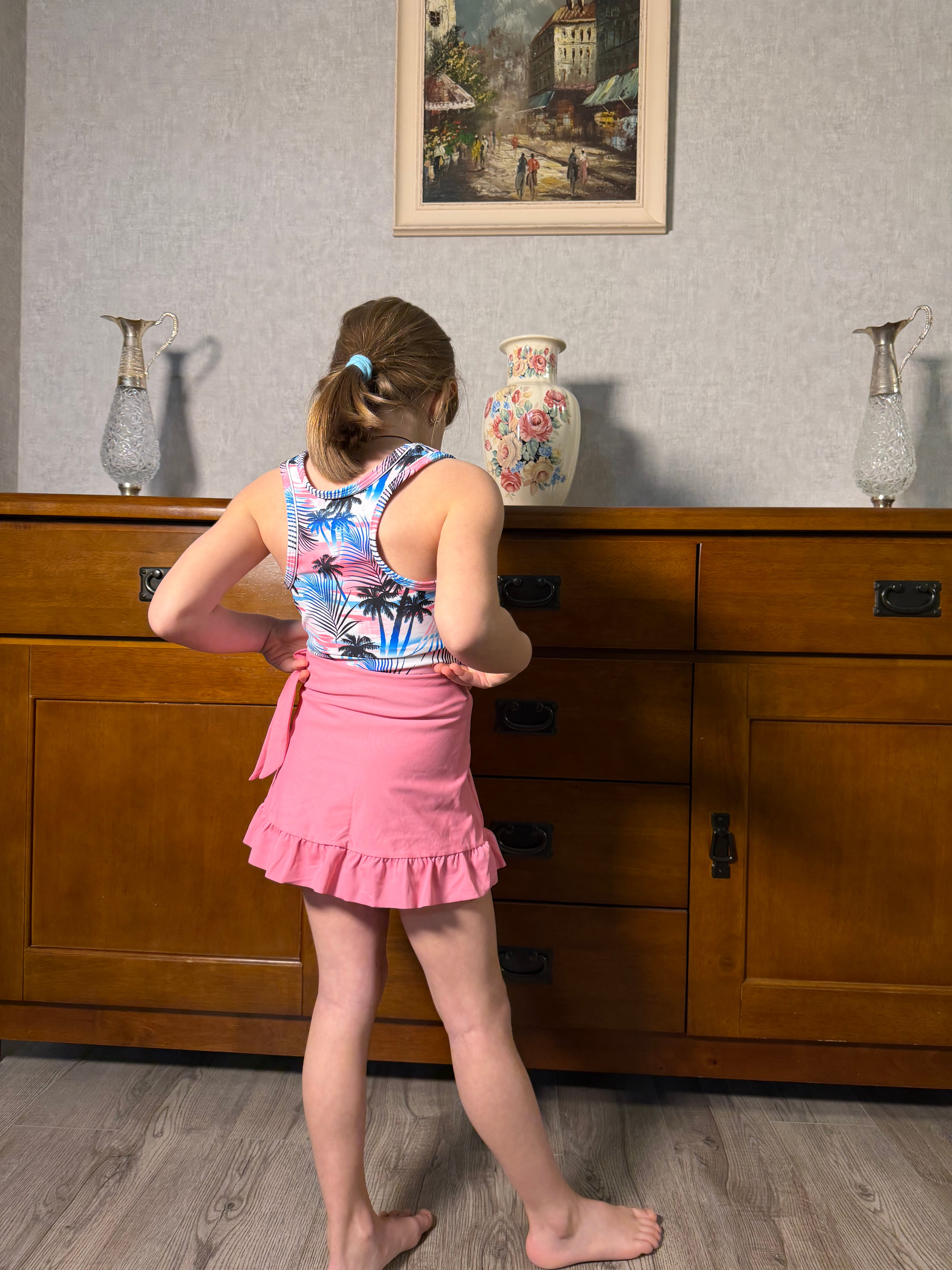 Back view of a girl wearing a pink palm tree 3 piece tankini swim set, showing the printed swim top, bottoms and ruffle skirt detail.