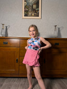 Girl wearing a pink palm tree 3 piece tankini swimsuit set indoors, showing the ruffle swim top, matching bottoms and swim skirt.