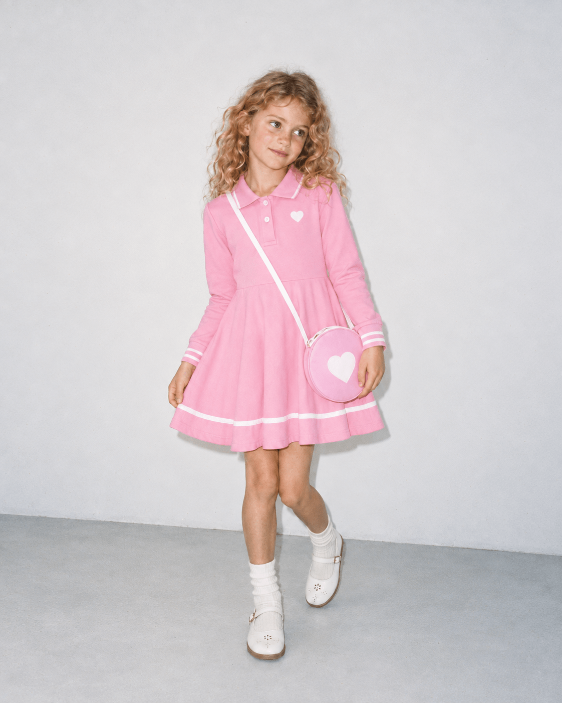 Girl wearing a pink long sleeve polo dress with heart shoulder bag in a front standing pose, showing the flared skirt and contrast trim.