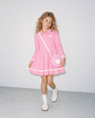 Girl wearing a pink long sleeve polo dress with heart shoulder bag in a front standing pose, showing the flared skirt and contrast trim.