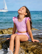 Girl wearing a pink striped ruffle bikini set while sitting by the ocean, showing a cute summer swimsuit for beach vacations.