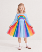 Girl wearing a rainbow tulle dress while holding the layered skirt, showing the colorful rainbow tulle layers, long sleeve design and princess-style silhouette.