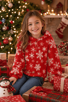 Girl wearing a red snowflake fleece hoodie beside Christmas gifts and a decorated tree, showing a festive holiday outfit for winter celebrations.