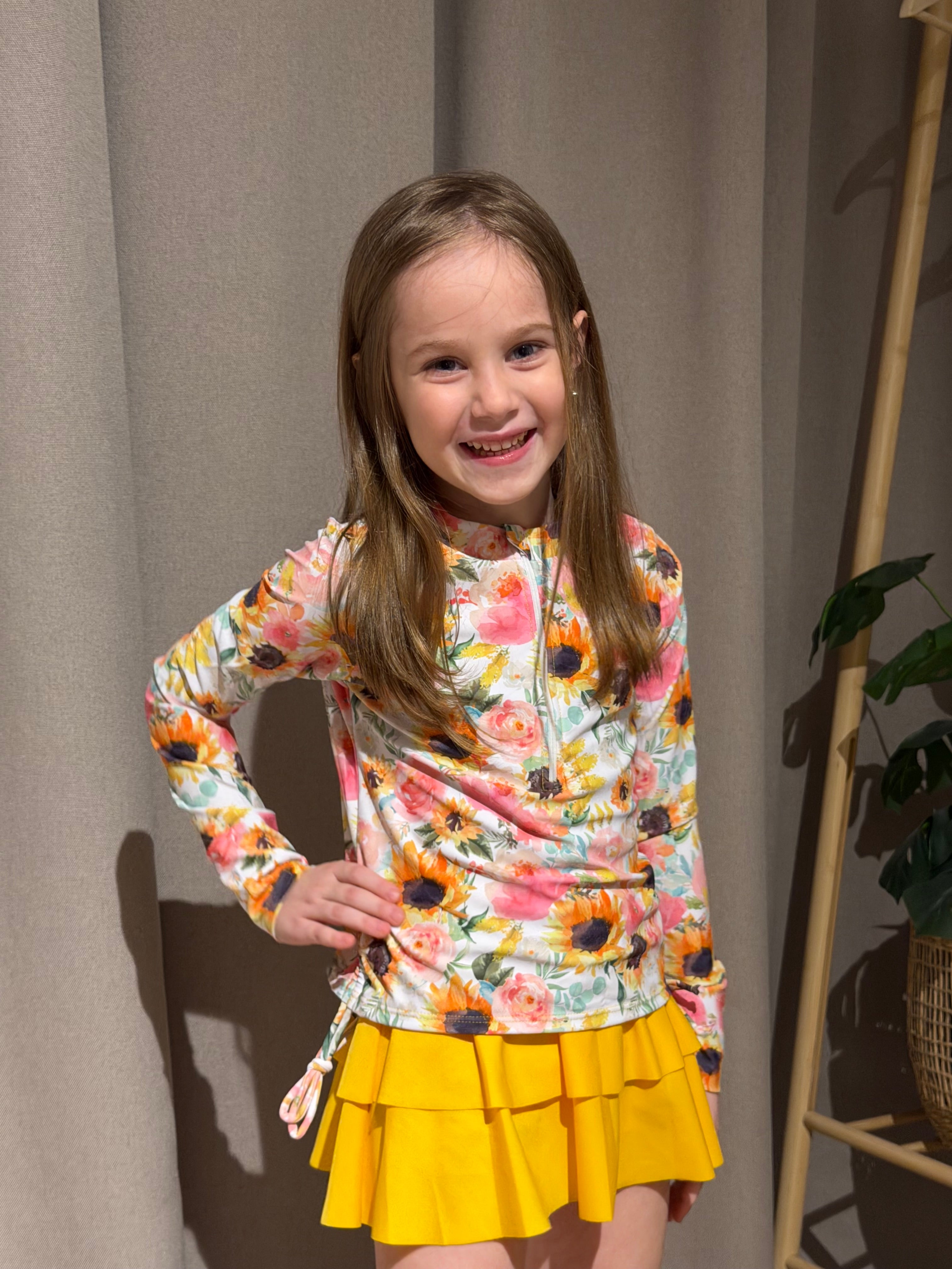 Girl wearing a Yellow Sunflowers rash guard swimsuit set indoors, showing the colorful sunflower print, long sleeve top and matching swim bottoms.