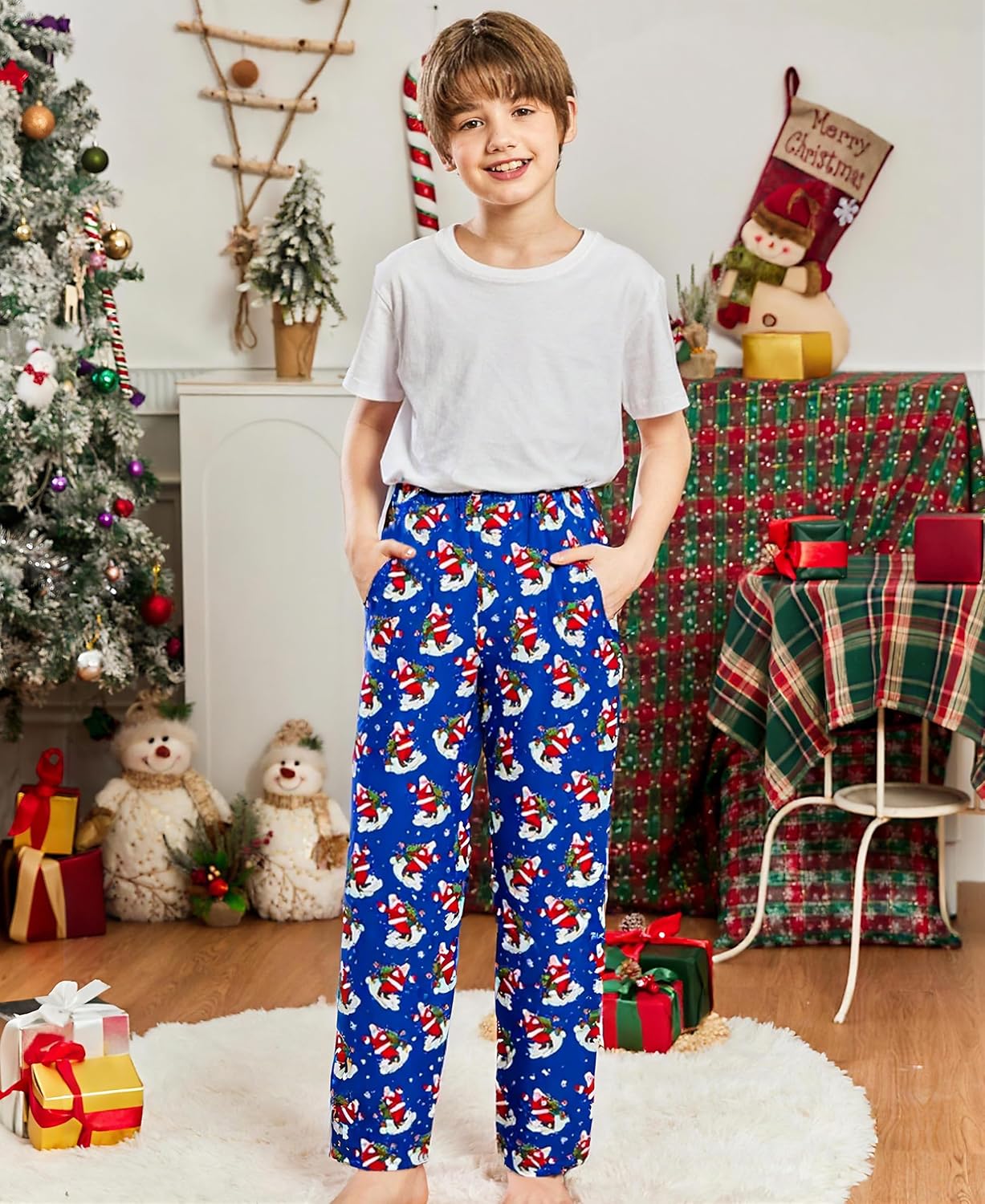 Child wearing blue Santa Claus pajama pants indoors beside Christmas decorations, showing the relaxed fit and festive holiday print.