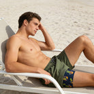 Man relaxing on the beach wearing green 2-in-1 compression liner swim trunks with tropical print inner layer