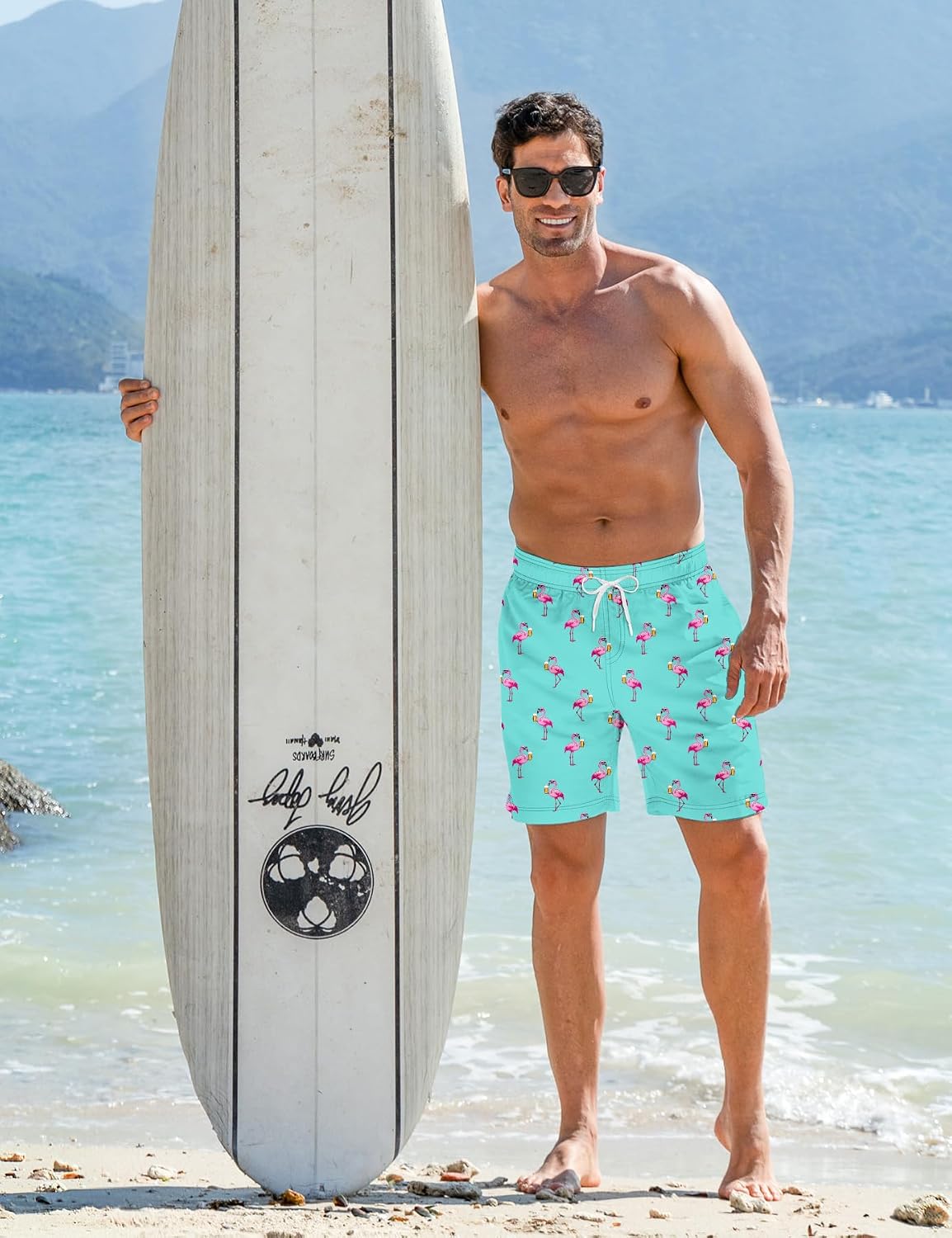 Man wearing blue flamingo swim trunks while standing beside a surfboard on the beach, styled for swimming, surfing and summer vacations.