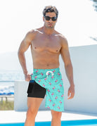 Front view of a man wearing blue flamingo swim trunks with one side lifted to reveal the built-in black compression liner.