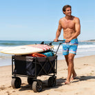 Man wearing blue geometric print swim trunks while pulling a beach wagon with surf gear along the shoreline.