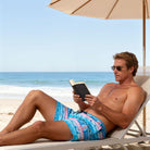 Man relaxing under a beach umbrella while wearing blue geometric print swim trunks, styled for comfortable seaside leisure.