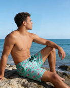 Man sitting on seaside rocks wearing green flamingo swim trunks, styled for relaxed beach and vacation wear.