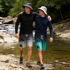 Men wearing 2-in-1 swim trunks while walking beside a rocky stream, showing a versatile summer outfit for hiking and outdoor activities.