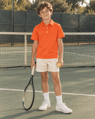 Boy wearing an orange polo shirt on a tennis court, showing the classic collar, short sleeves and relaxed sporty outfit style.