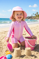 Toddler girl wearing a pink striped full coverage rash guard swimsuit with matching sun hat during an outdoor summer activity.
