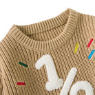 Close-up of toddler khaki knit sweater showing the ribbed crew neckline, raised white 1/2 design and colorful embroidered accents.