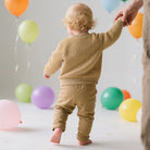 Back view of a toddler wearing a khaki knit birthday sweater while standing beside colorful balloons at a celebration.