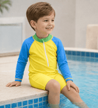 Toddler boy wearing a yellow colorblock rash guard swimsuit while sitting by the pool, showing the long sleeve one-piece swimwear fit.