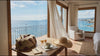 A sunlit seaside room with ocean views, flowing curtains, a travel bag, coffee and a straw hat.